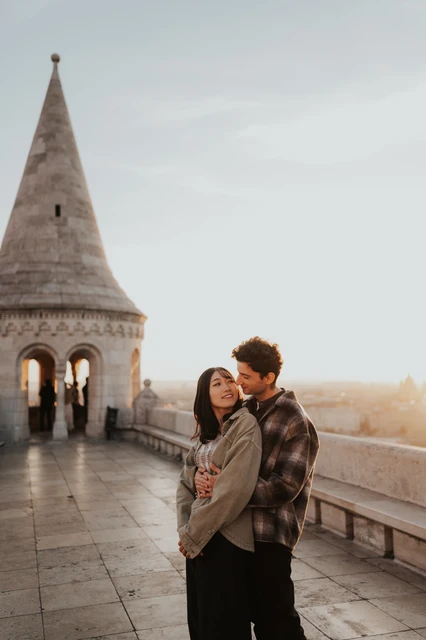 Budapest engagement photoshoot: a shared smile above the city The couple smiling in an embrace on the walkway at Fisherman’s Bastion, with the city visible beyond the stone wall during their Budapest engagement photoshoot.