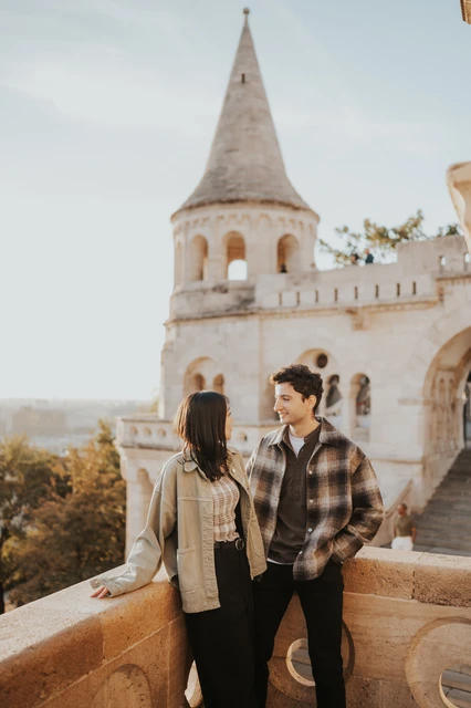 Budapest engagement photoshoot: a shared smile beside the terrace wall The couple smiling at one another beside the stone terrace wall, with a Bastion tower rising behind them during their Budapest engagement photoshoot.