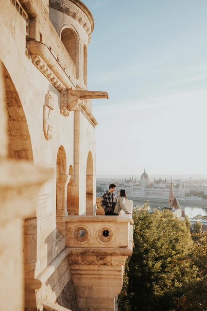 Budapest engagement photoshoot: a sunlit balcony overlooking Parliament A wide vertical view of the couple on a sunlit Bastion balcony, with trees, the Danube and Parliament beyond them during their Budapest engagement photoshoot.