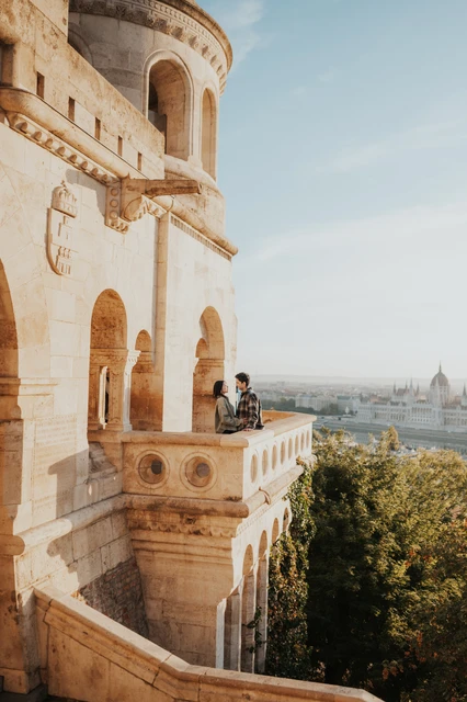 Budapest engagement photoshoot: a wide portrait framed by the tower façade The couple on the small Bastion balcony, with the tall stone façade to one side and the sunlit Budapest skyline beyond during their Budapest engagement photoshoot.