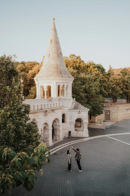 Budapest engagement photoshoot: a wide view of the lower Bastion tower An elevated view of the couple near a lower Bastion tower, surrounded by pale stone architecture, trees and open paving during their Budapest engagement photoshoot.