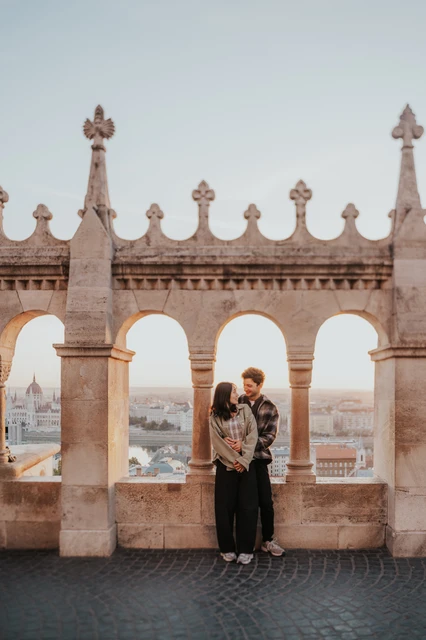 Budapest engagement photoshoot: an embrace framed by the Bastion arcade The couple embracing beneath the central arch at Fisherman’s Bastion, with the ornate stone arcade above them during their Budapest engagement photoshoot.