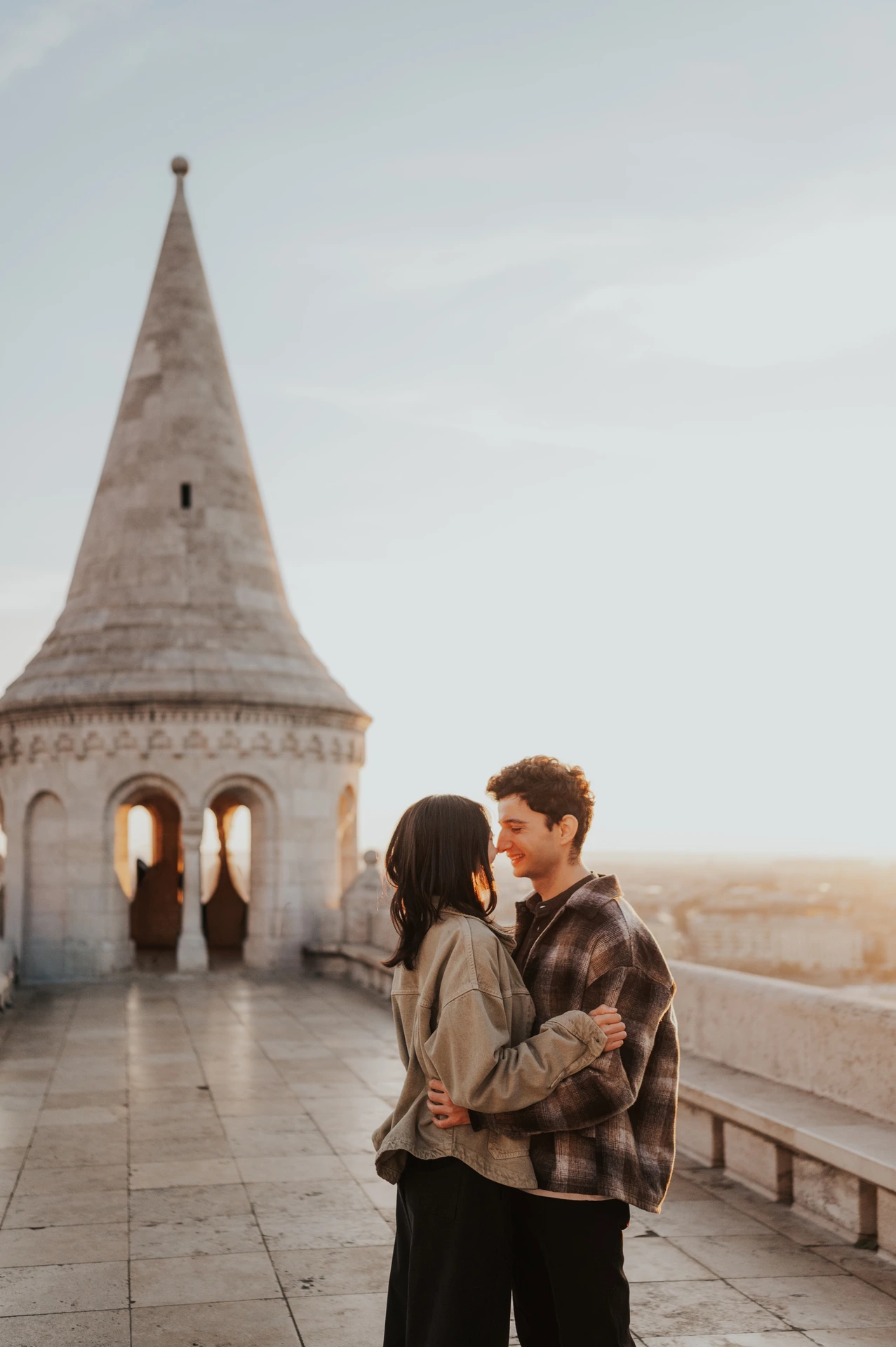 The couple embracing in warm early morning light beside the stone balustrade, with the Bastion tower rising behind them during their Budapest engagement photoshoot.