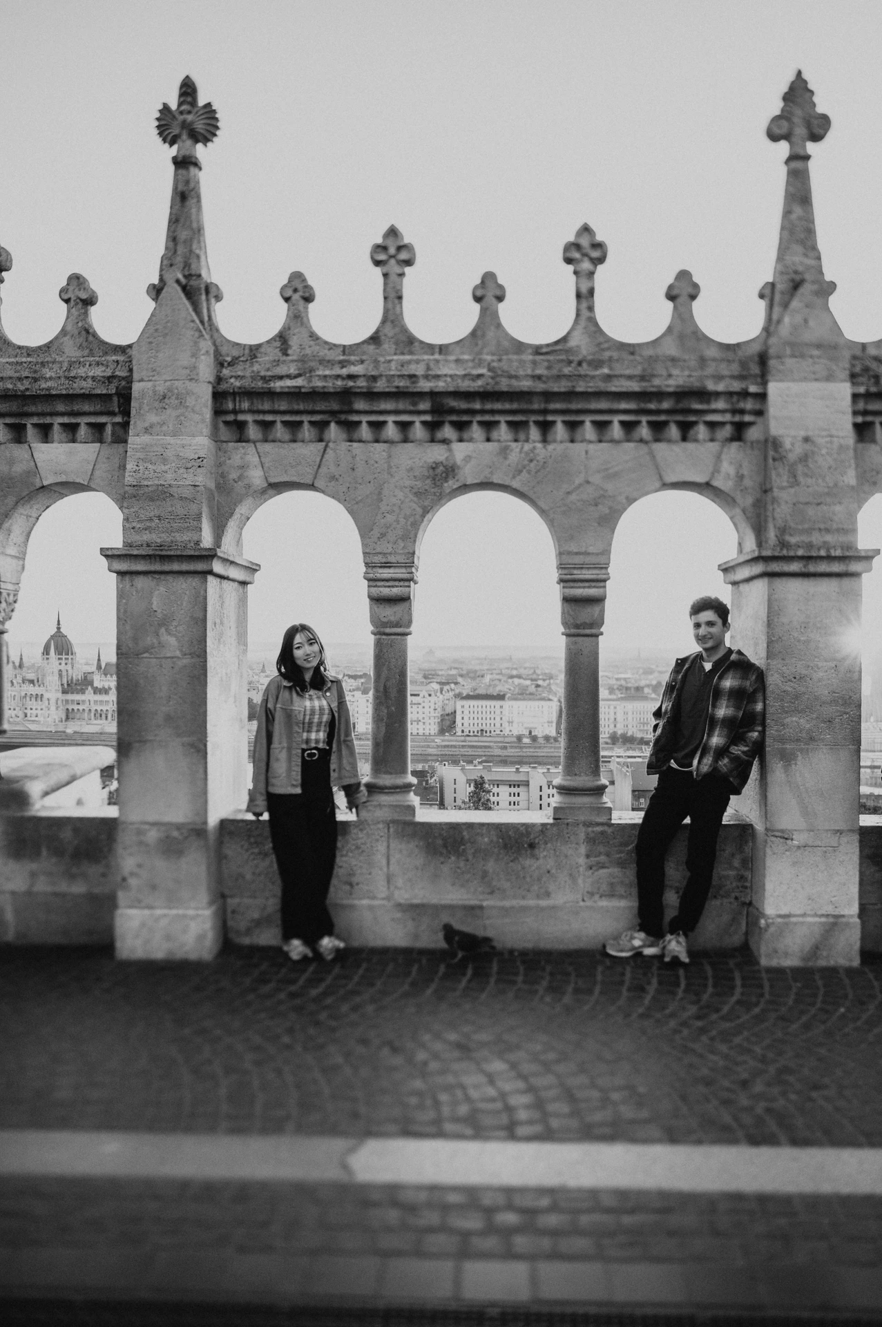 Black-and-white view of the couple beneath separate arches at Fisherman’s Bastion, with their casual outfits and trainers in full view during their Budapest engagement photoshoot.