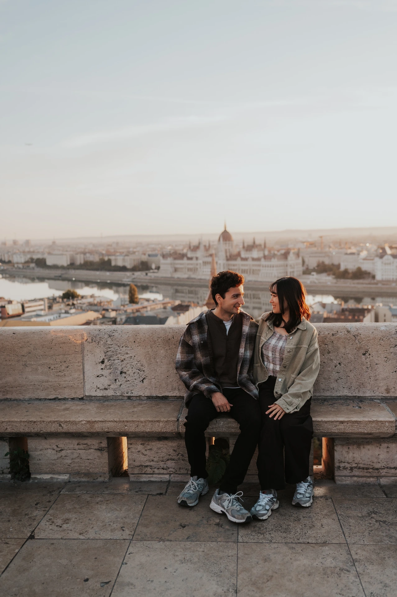 The couple seated closely on a stone bench at Fisherman’s Bastion, with their trainers visible and the city beyond the terrace during their Budapest engagement photoshoot.