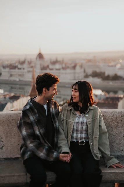 Budapest engagement photoshoot: close smiles with Parliament in the backdrop A close portrait of the couple smiling at one another on the stone bench, with the Hungarian Parliament softly blurred behind them during their Budapest engagement photoshoot.