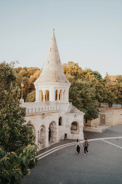 Budapest engagement photoshoot: holding hands below the pale stone tower The couple holding hands on the open paving below a Bastion tower, seen from an elevated viewpoint during their Budapest engagement photoshoot.