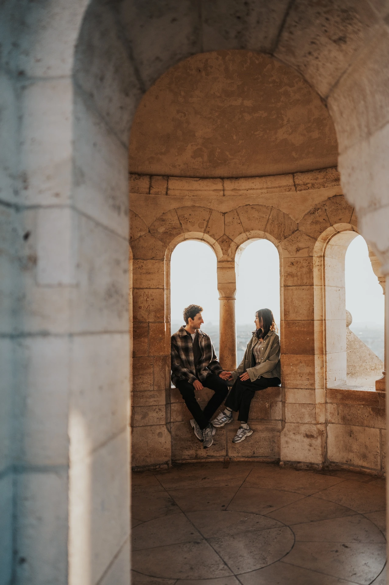 The couple holding hands while seated inside a sheltered stone tower, framed by its arched openings and vaulted ceiling during their Budapest engagement photoshoot.