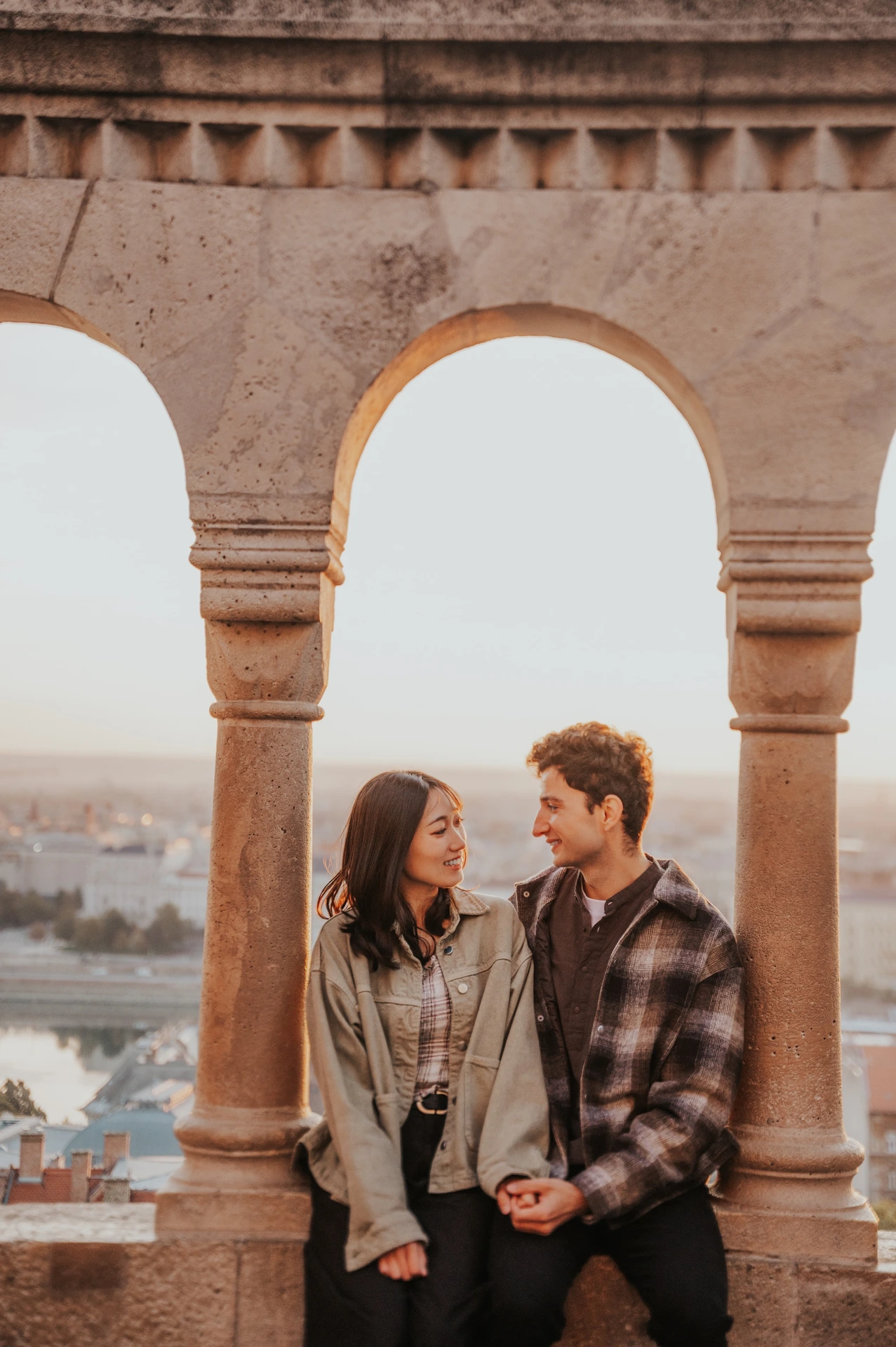 The couple seated together on a stone ledge beneath a Bastion arch, holding hands and looking towards each other during their Budapest engagement photoshoot.