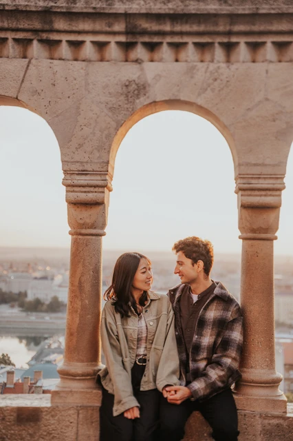 The couple seated together on a stone ledge beneath a Bastion arch, holding hands and looking towards each other during their Budapest engagement photoshoot.