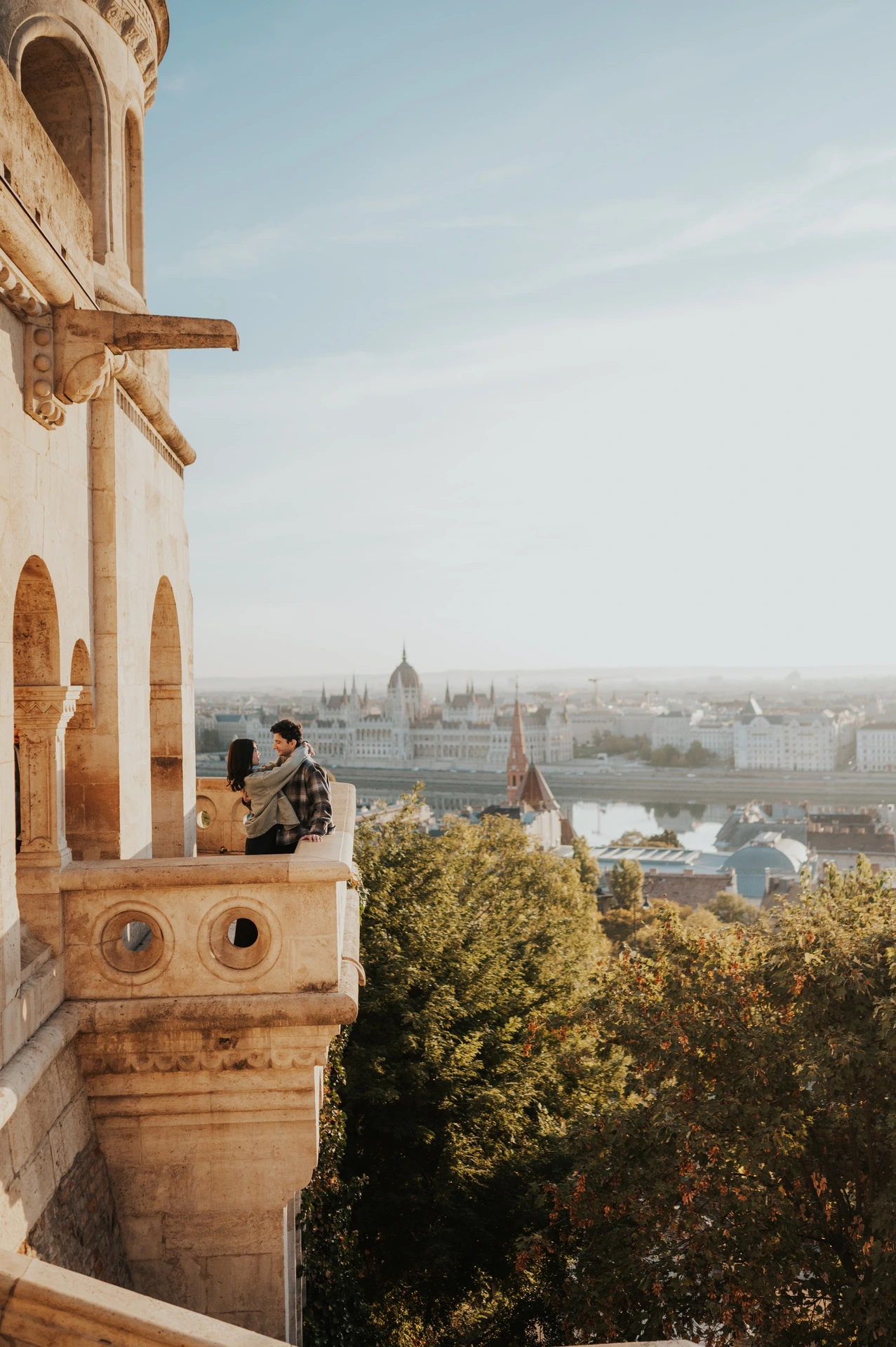 The couple leaning together on the stone balcony wall at Fisherman’s Bastion, overlooking the trees and river during their Budapest engagement photoshoot.