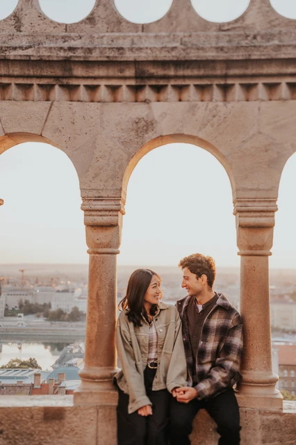 Budapest engagement photoshoot: shared looks framed by a stone arch The couple looking at each other on the stone ledge, framed by the pale arches at Fisherman’s Bastion during their Budapest engagement photoshoot.