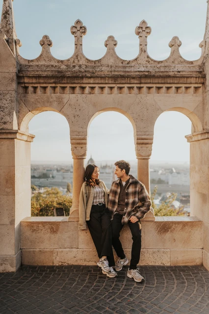 Budapest engagement photoshoot: sharing a look beneath the morning-lit arcade The couple looking towards one another beneath the pale stone arches at Fisherman’s Bastion, with warm morning light behind them during their Budapest engagement photoshoot.