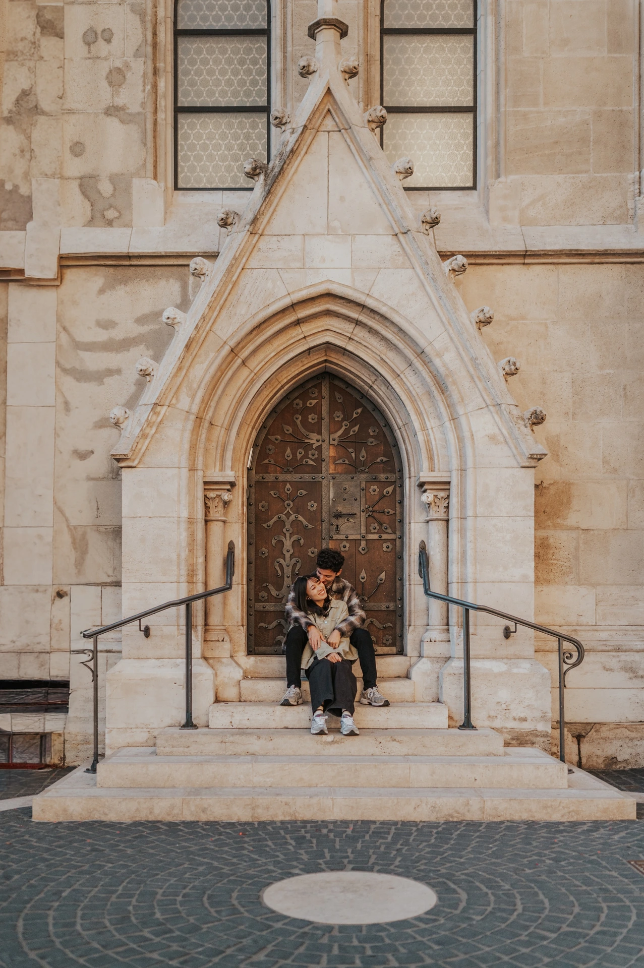 The couple seated closely together beneath the ornate wooden door, with the stone arch and railings framing the steps during their Budapest engagement photoshoot.