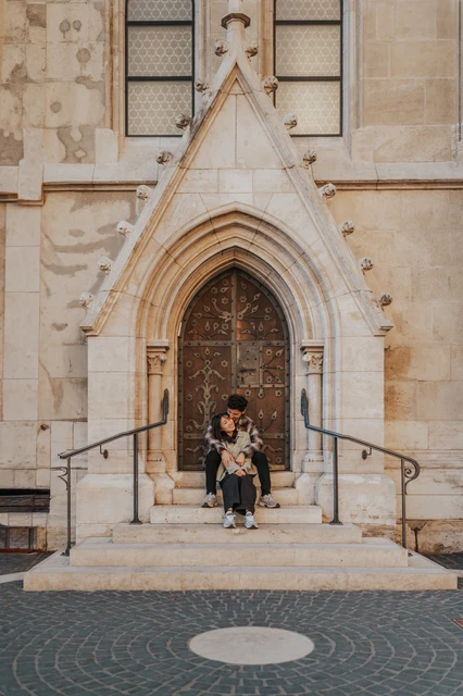 Budapest engagement photoshoot: sitting together beneath the wooden door The couple seated closely together beneath the ornate wooden door, with the stone arch and railings framing the steps during their Budapest engagement photoshoot.