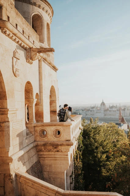 Budapest engagement photoshoot: standing together on the corner balcony The couple standing close at the corner of the sunlit Bastion balcony, framed by the tower wall and city views during their Budapest engagement photoshoot.