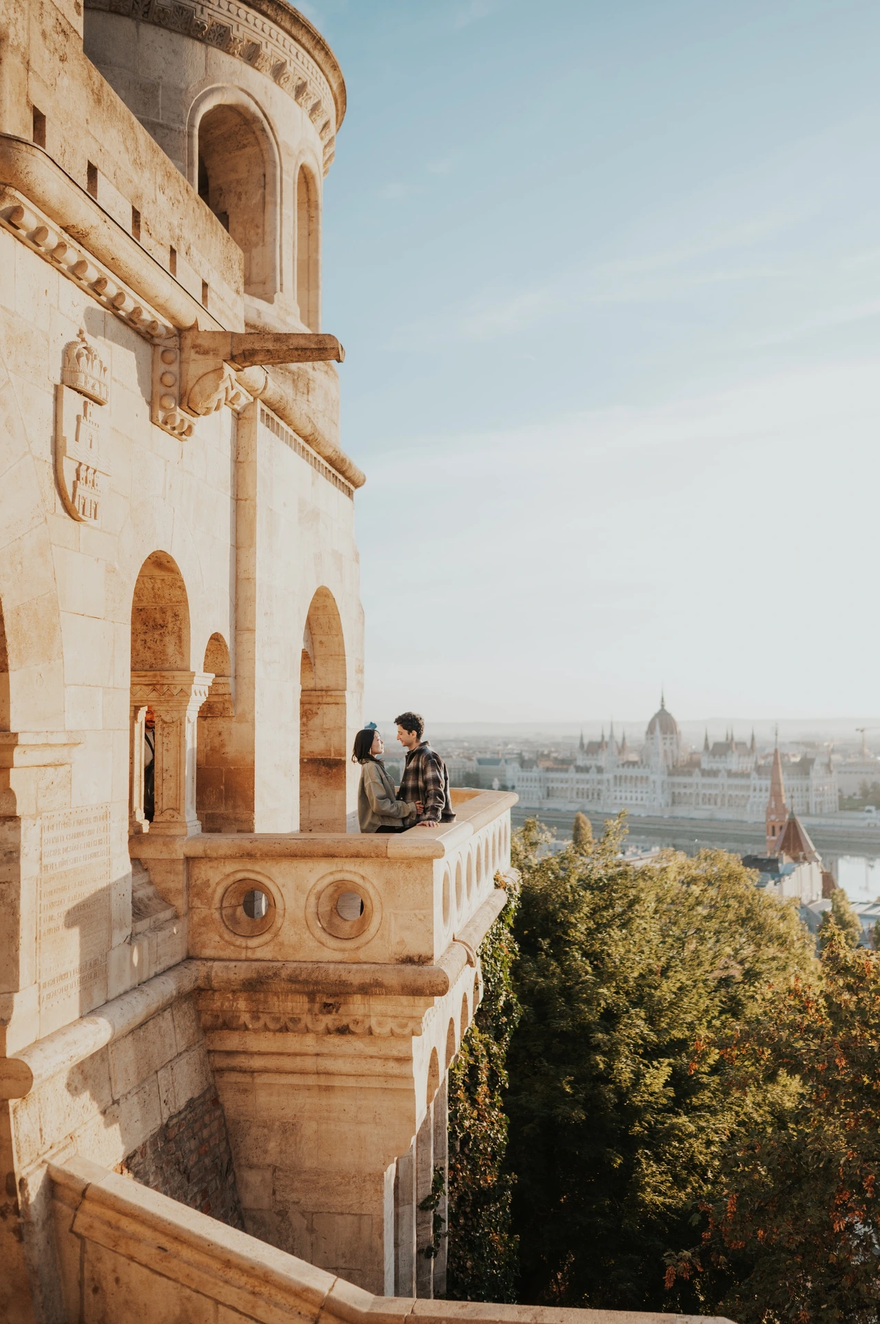 The couple leaning close together against the balcony balustrade at Fisherman’s Bastion, with the city across the river behind them during their Budapest engagement photoshoot.