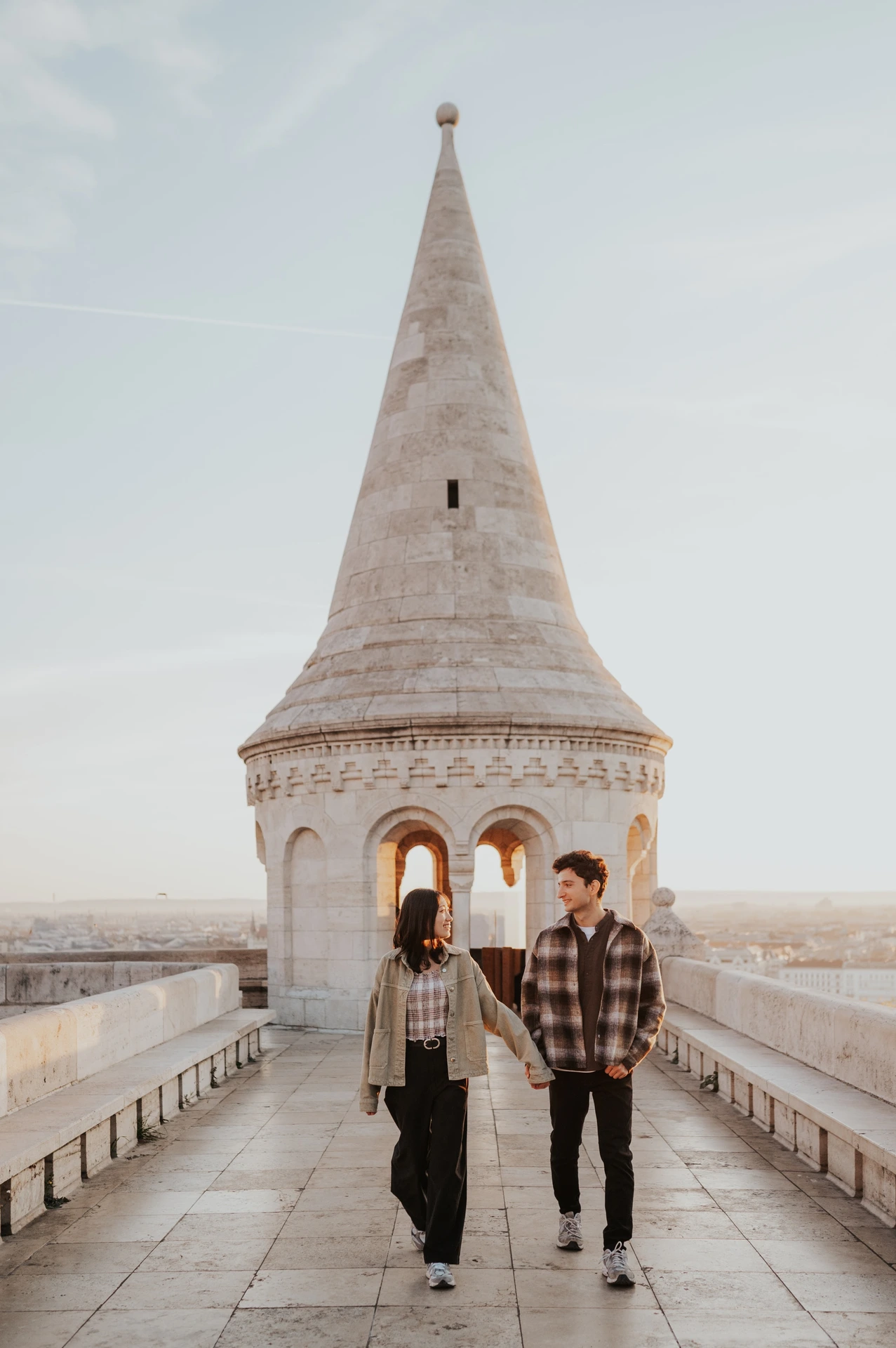 A full-length view of the couple walking together in trainers, with the conical Bastion tower behind them during their Budapest engagement photoshoot.