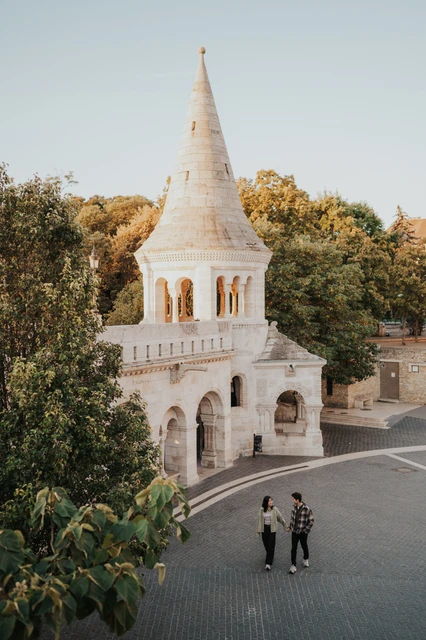 Budapest engagement photoshoot: two figures beneath the tower and trees The couple standing close on the lower terrace, with the pointed Bastion tower and leafy trees filling the wider view during their Budapest engagement photoshoot.