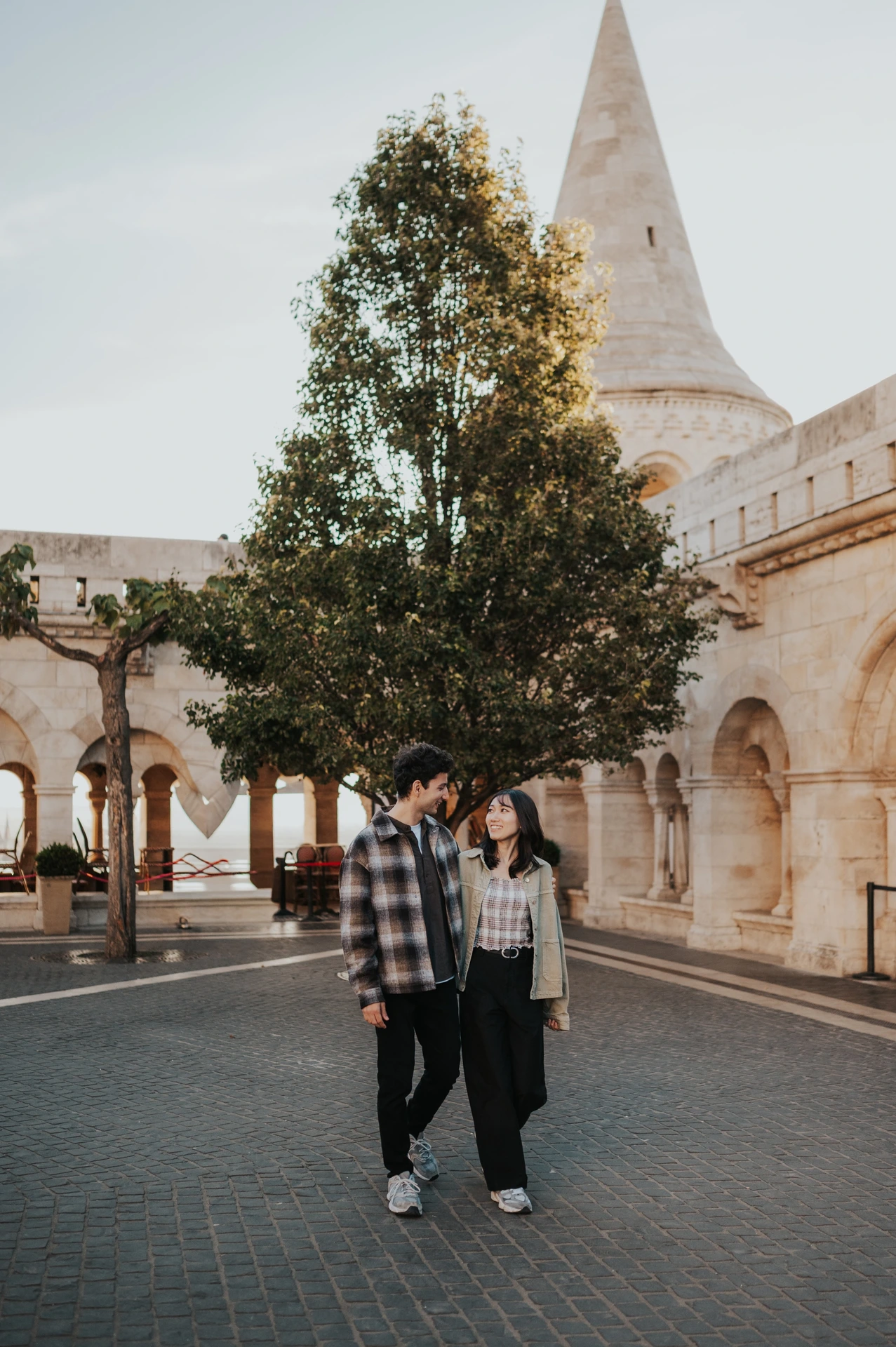 The couple walking hand in hand across the courtyard at Fisherman’s Bastion, with a tree and the stone arcade behind them during their Budapest engagement photoshoot.