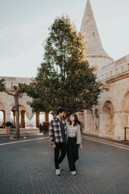 The couple walking hand in hand across the courtyard at Fisherman’s Bastion, with a tree and the stone arcade behind them during their Budapest engagement photoshoot.