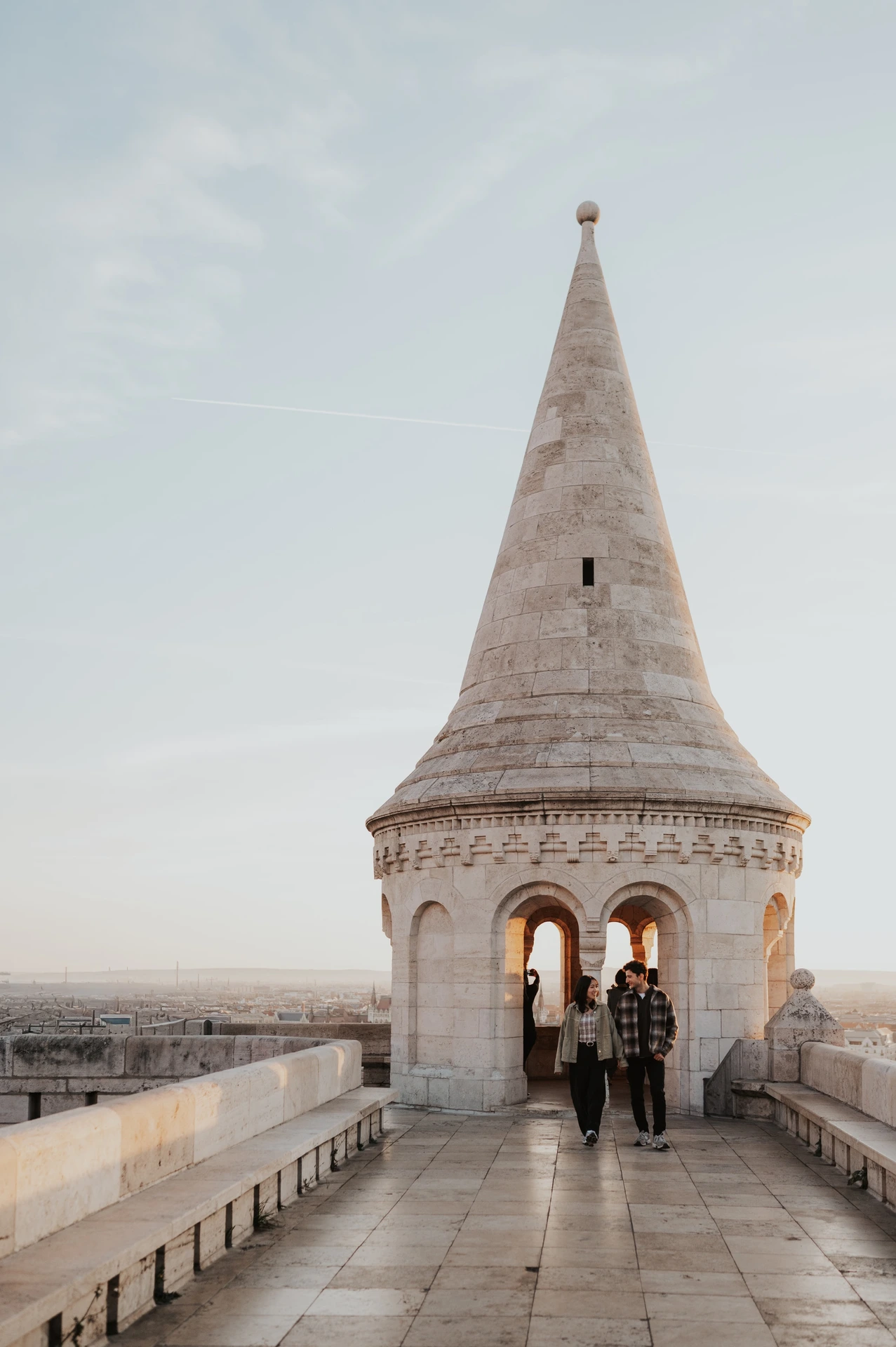 A distant view of the couple holding hands on the stone walkway towards the pointed tower at Fisherman’s Bastion during their Budapest engagement photoshoot.