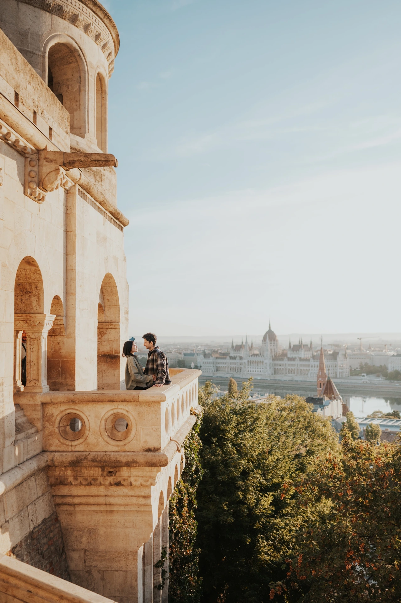 A wide view of the couple embracing on the balcony, with warm sunlight across the Bastion’s carved stone façade during their Budapest engagement photoshoot.