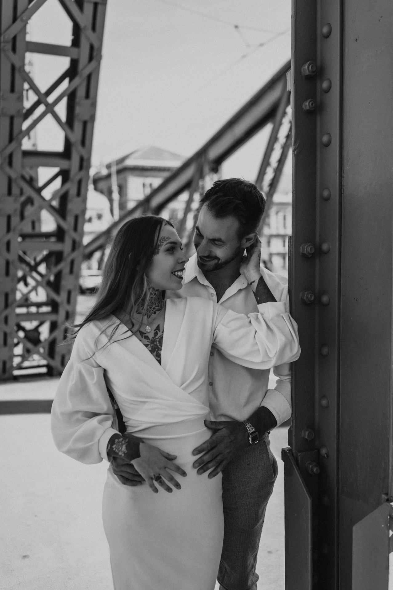 Black-and-white photograph of the couple embracing beside the riveted steelwork of Liberty Bridge during their engagement photoshoot in Budapest.