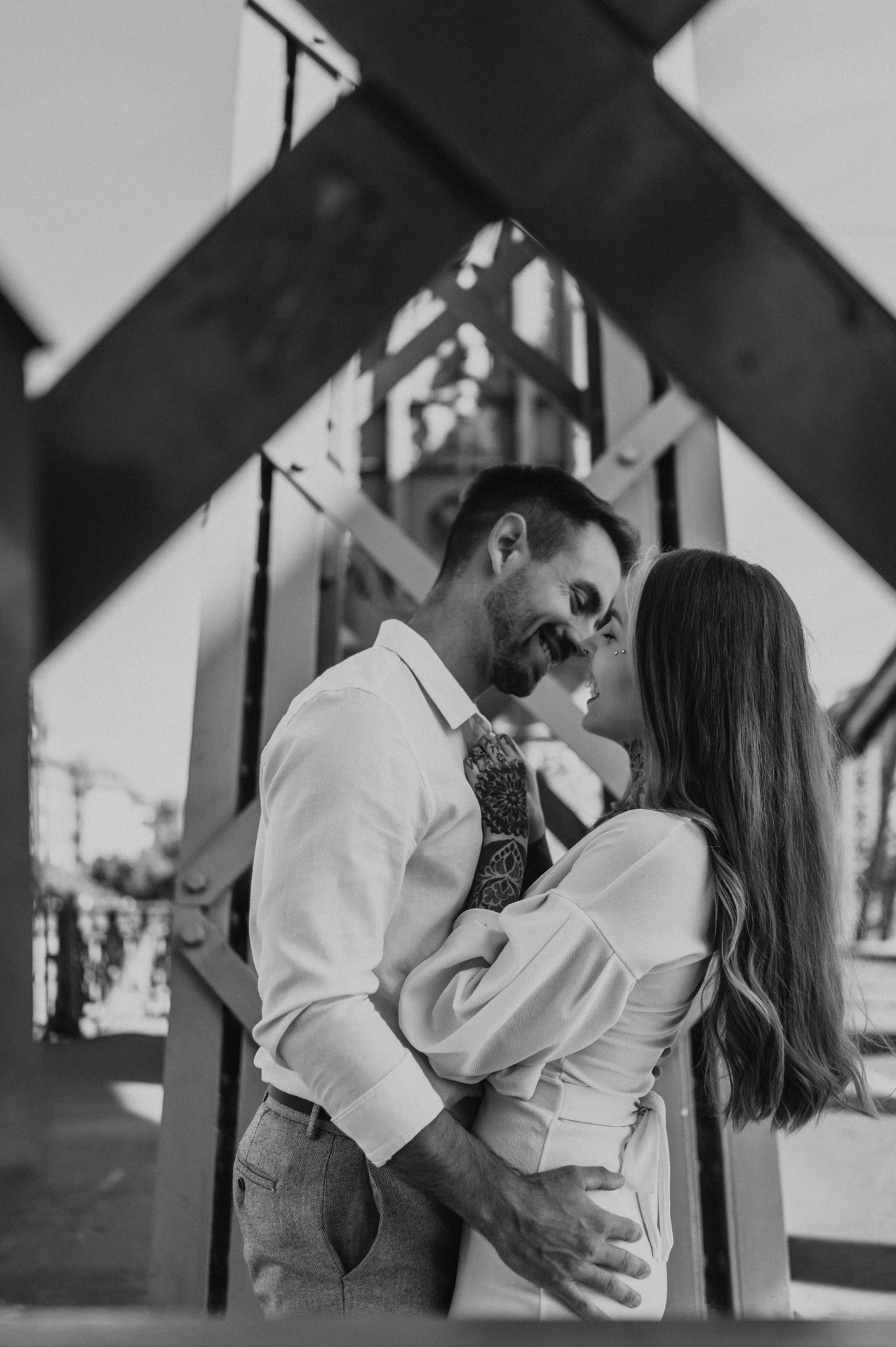 Black-and-white photograph of the couple embracing beneath the diagonal steel beams of Liberty Bridge during their engagement photoshoot in Budapest.