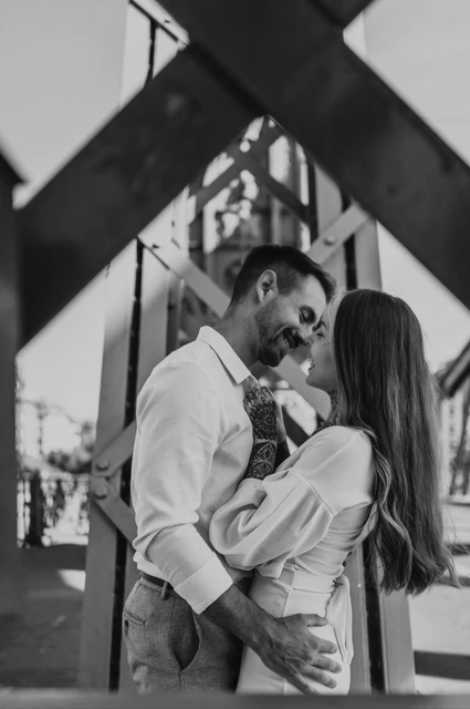 An embrace among the steel beams in black and white | Budapest engagement photoshoot Black-and-white photograph of the couple embracing beneath the diagonal steel beams of Liberty Bridge during their engagement photoshoot in Budapest.