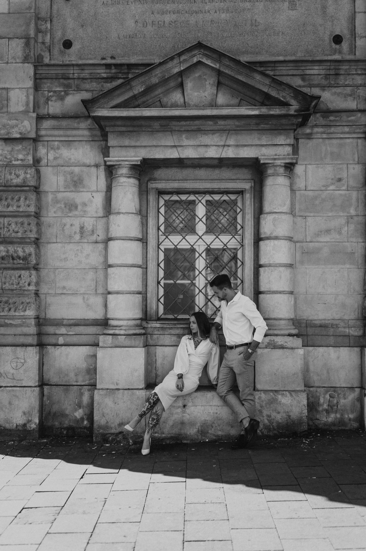 Black-and-white photograph of the couple leaning together beside a carved stone doorway, with the woman sitting on a low ledge during their engagement photoshoot in Budapest.