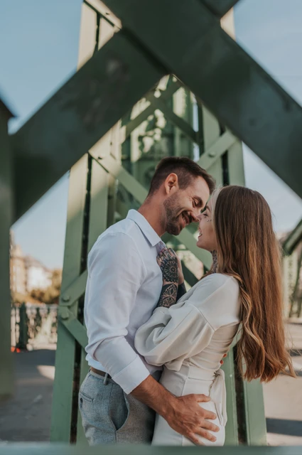 An embrace beneath the diagonal green steelwork | Budapest engagement photoshoot The couple embracing beneath the diagonal green steelwork of Liberty Bridge, with tattoo details visible during their engagement photoshoot in Budapest.