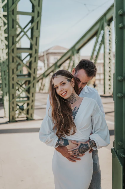 A smiling portrait among the bridge steelwork | Budapest engagement photoshoot The woman smiling towards the camera as her partner embraces her beside the green steelwork of Liberty Bridge during their engagement photoshoot in Budapest.