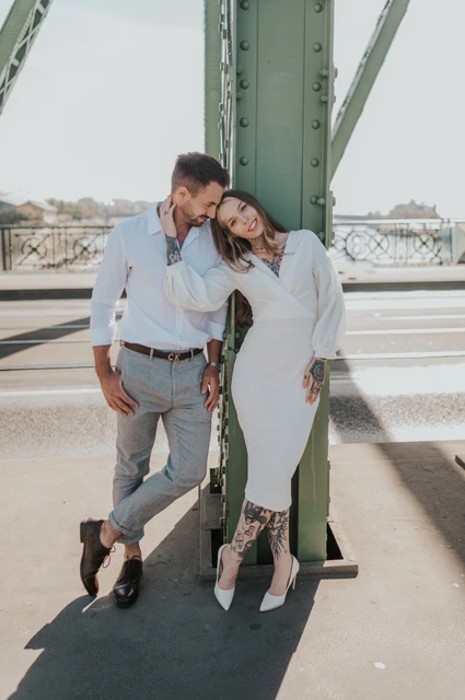 Together against a green bridge pillar | Budapest engagement photoshoot The couple leaning together against a green steel pillar on Liberty Bridge, with their full white outfits visible during their engagement photoshoot in Budapest.