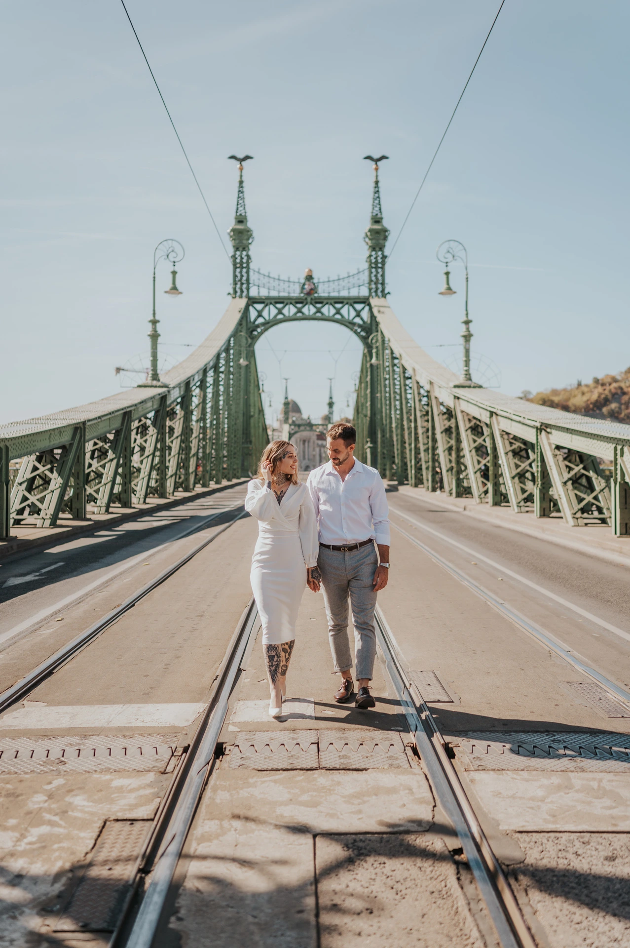 The couple holding hands with the green towers, lamps and long perspective of Liberty Bridge behind them during their engagement photoshoot in Budapest.