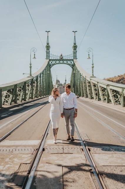 Hand in hand with the green bridge beyond | Budapest engagement photoshoot The couple holding hands with the green towers, lamps and long perspective of Liberty Bridge behind them during their engagement photoshoot in Budapest.