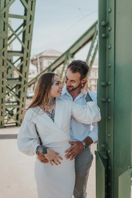 An embrace beside the riveted green pillar | Budapest engagement photoshoot The couple looking at one another while embracing beside a riveted green pillar on Liberty Bridge during their engagement photoshoot in Budapest.