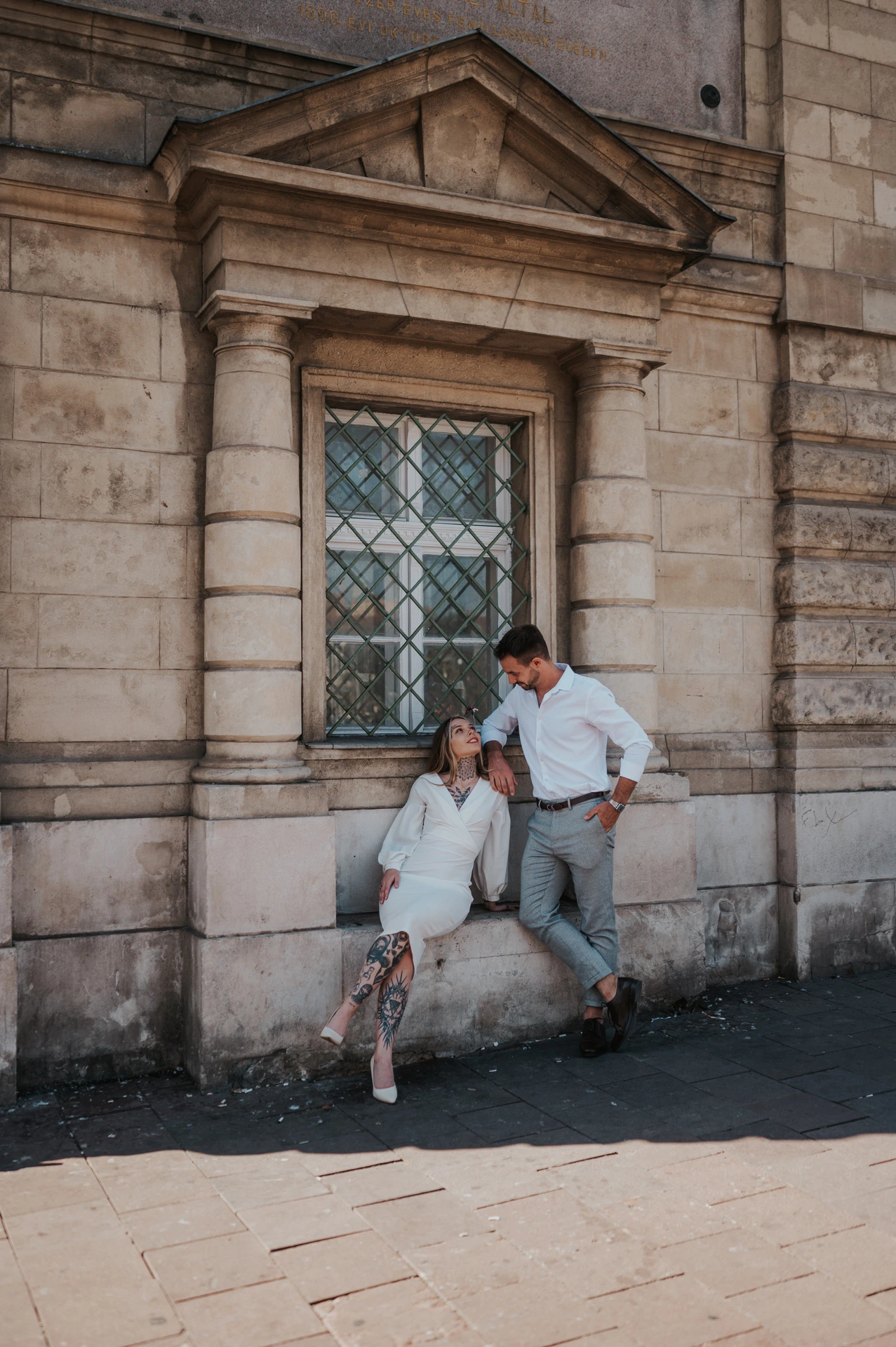 The couple looking towards the camera beside a wooden doorway set into a pale stone building during their engagement photoshoot in Budapest.