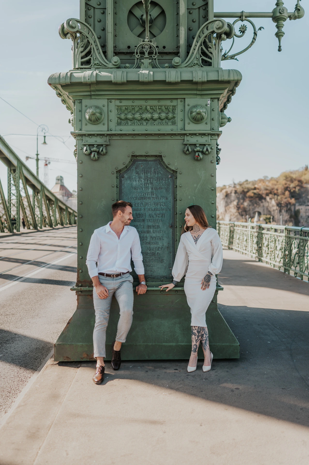 The couple standing on opposite sides of a large ornate green lamp-post base on Liberty Bridge during their engagement photoshoot in Budapest.