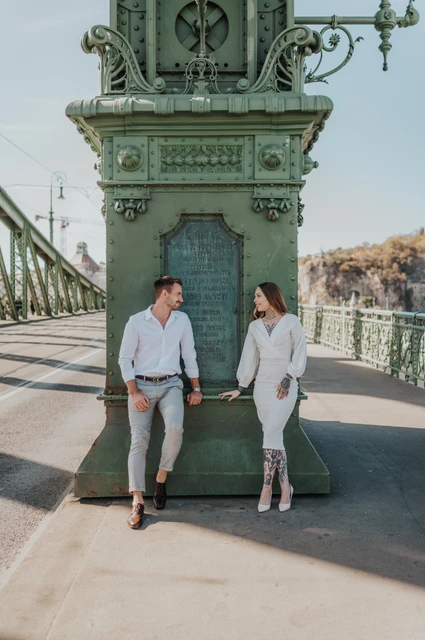The couple standing on opposite sides of a large ornate green lamp-post base on Liberty Bridge during their engagement photoshoot in Budapest.