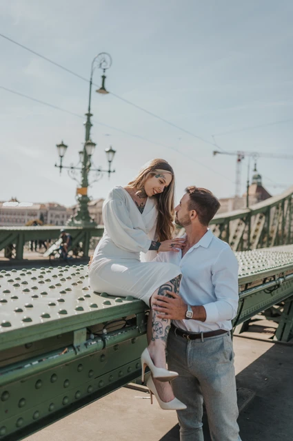 A relaxed portrait beside the green bridge railing | Budapest engagement photoshoot The woman sitting on a low green section of Liberty Bridge while her partner stands close beside her during their engagement photoshoot in Budapest.