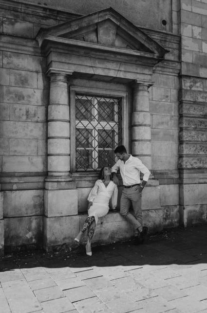 Together by the carved wall in black and white | Budapest engagement photoshoot Black-and-white photograph of the couple sitting and standing close together beside a wooden doorway and carved stone wall during their engagement photoshoot in Budapest.