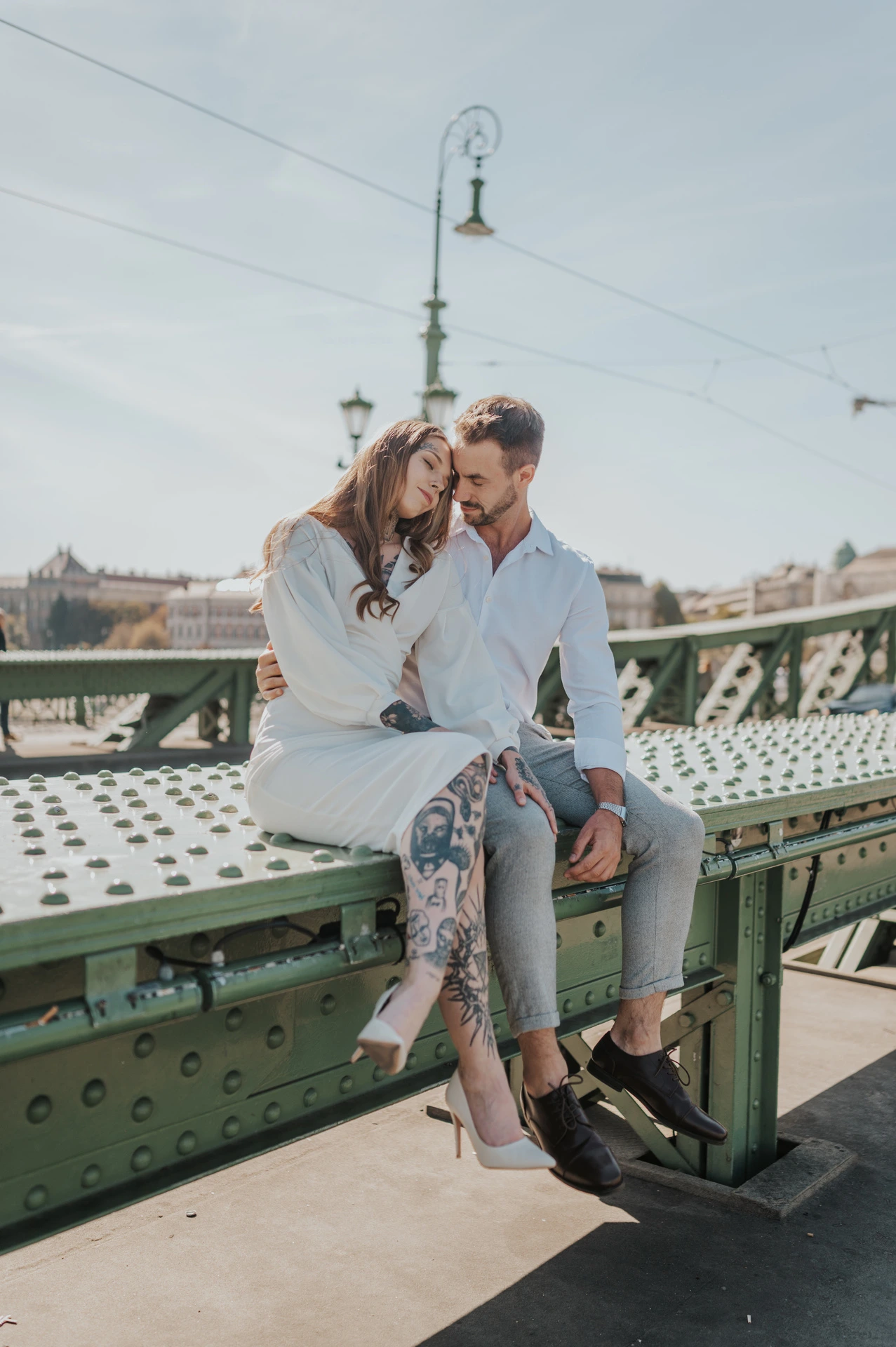 The couple sitting close together on a low green section of Liberty Bridge with the bridge lamps behind them during their engagement photoshoot in Budapest.