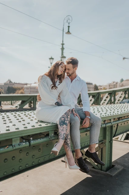 Sitting together among the green bridge details | Budapest engagement photoshoot The couple sitting close together on a low green section of Liberty Bridge with the bridge lamps behind them during their engagement photoshoot in Budapest.