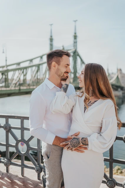 Smiles beside the river and bridge | Budapest engagement photoshoot The couple smiling towards one another beside the riverside railing with Liberty Bridge in the background during their engagement photoshoot in Budapest.