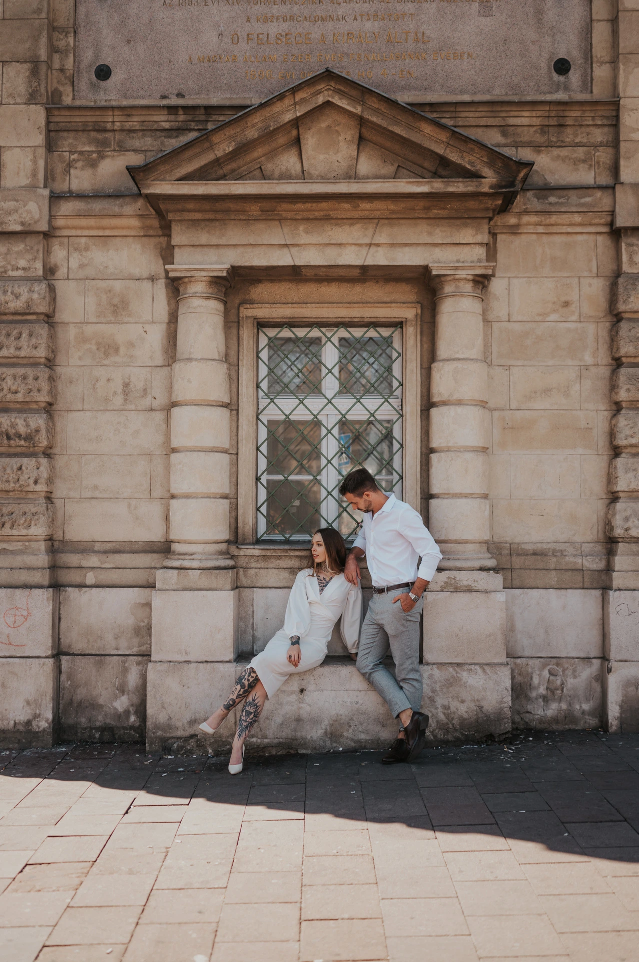 The woman sitting on a stone ledge with her partner leaning close beside her in front of a wooden doorway during their engagement photoshoot in Budapest.