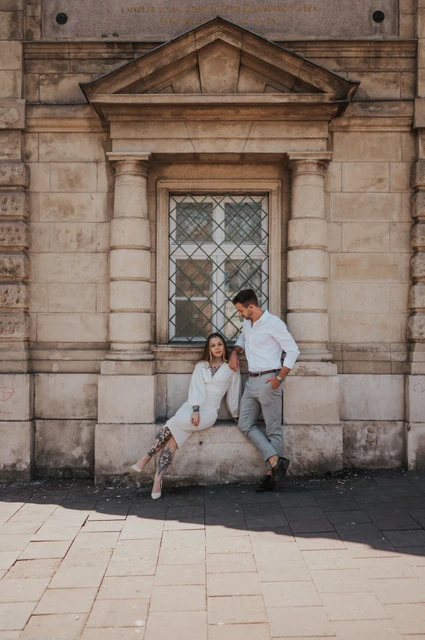 A portrait beside the carved stone doorway | Budapest engagement photoshoot The woman sitting on a stone ledge beside her partner in front of a wooden doorway and carved stone building during their engagement photoshoot in Budapest.