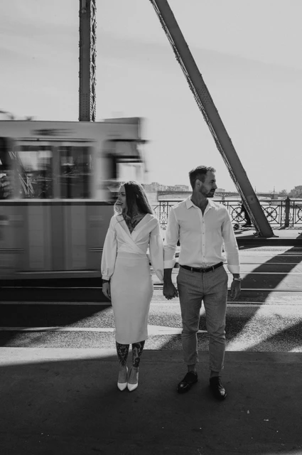 A bridge walk and tram in black and white | Budapest engagement photoshoot Black-and-white photograph of the couple walking hand in hand on Liberty Bridge with a tram passing behind them during their engagement photoshoot in Budapest.