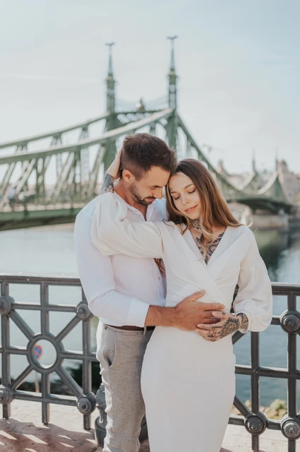 White outfits beside the Danube | Budapest engagement photoshoot The couple in white clothing looking down together beside the river, with the green bridge behind them during their engagement photoshoot in Budapest.