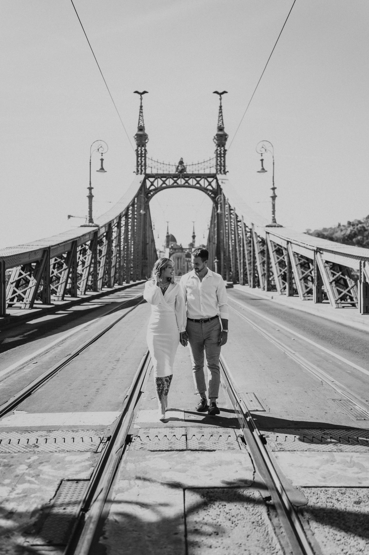 Black-and-white photograph of the couple holding hands with Liberty Bridge’s towers and railings stretching behind them during their engagement photoshoot in Budapest.